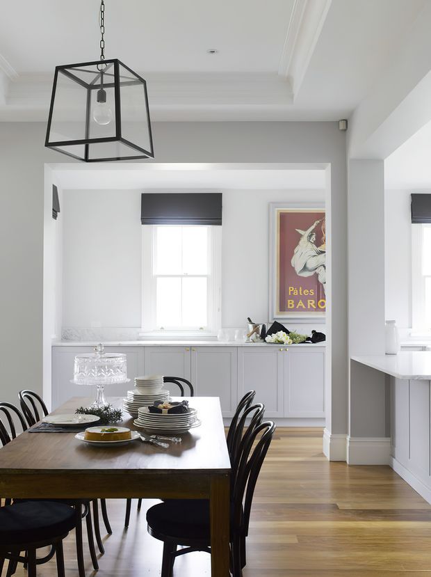 A bright, refined dining area with a dark timber dining table and curved black chairs in the foreground, opening into a white and pale-grey kitchen. The kitchen features traditional panelled cabinetry, a light benchtop, timber flooring, sash windows with dark roller blinds, and a framed vintage poster. A geometric black metal pendant hangs above the dining table, while decorative ceiling mouldings and substantial white trim add architectural character.