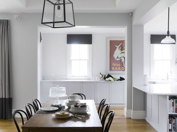 Bright, refined dining area in an open-plan home, featuring a substantial timber dining table with black bentwood-style chairs, set against white built-in cabinetry and a window seat or sideboard. The adjacent white kitchen is visible through a wide opening, with pendant lighting, pale walls, timber flooring, and a framed vintage-style poster. A large black-framed geometric pendant hangs above the dining table, complemented by a black pendant in the kitchen.