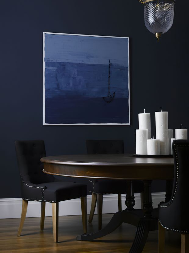 A moody, formal dining room with deep navy walls, a dark round pedestal dining table, and upholstered black dining chairs accented with nailhead trim. White pillar candles sit on the tabletop, while a blue abstract artwork and decorative pendant light add visual interest. Timber flooring and white skirting provide contrast.