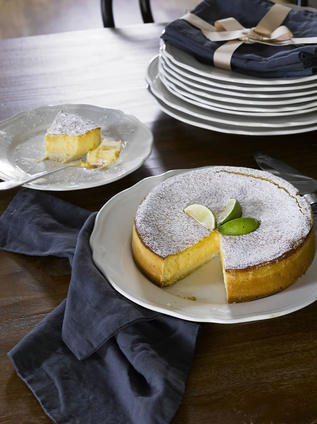 Close-up still life of a dining table set for dessert, featuring a large powdered-sugar cake garnished with lime slices on a white serving platter, a partially served slice on a plate, stacked plates with folded dark napkins, and a dark timber tabletop. The image is styled as a warm food and tablescape photograph.