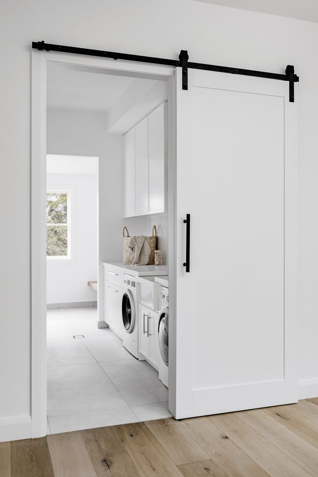Bright, white laundry room viewed through a wide barn-style sliding door. The room contains front-loading washer and dryer appliances beneath a pale benchtop, white shaker-style cabinetry, woven storage baskets, and large light-grey floor tiles. The foreground has light timber flooring, contrasting with the laundry tile floor, while black door hardware provides a strong accent.