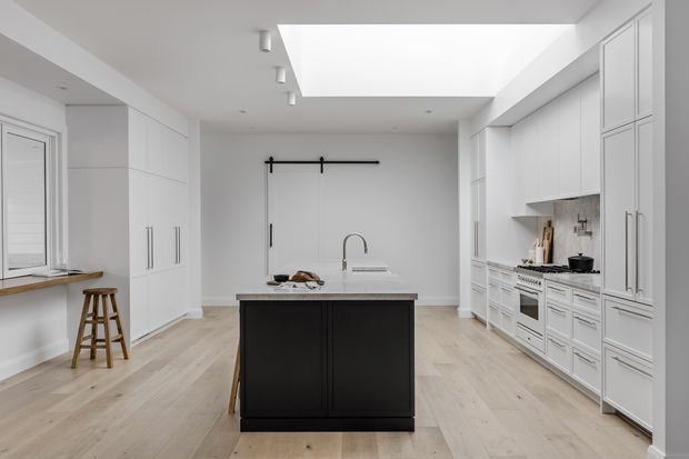 Bright, spacious contemporary kitchen with white floor-to-ceiling cabinetry, integrated appliances, pale timber flooring, and a contrasting dark island with a stone benchtop. A large skylight provides abundant natural light, while a small window bench and stools sit along the left wall.
