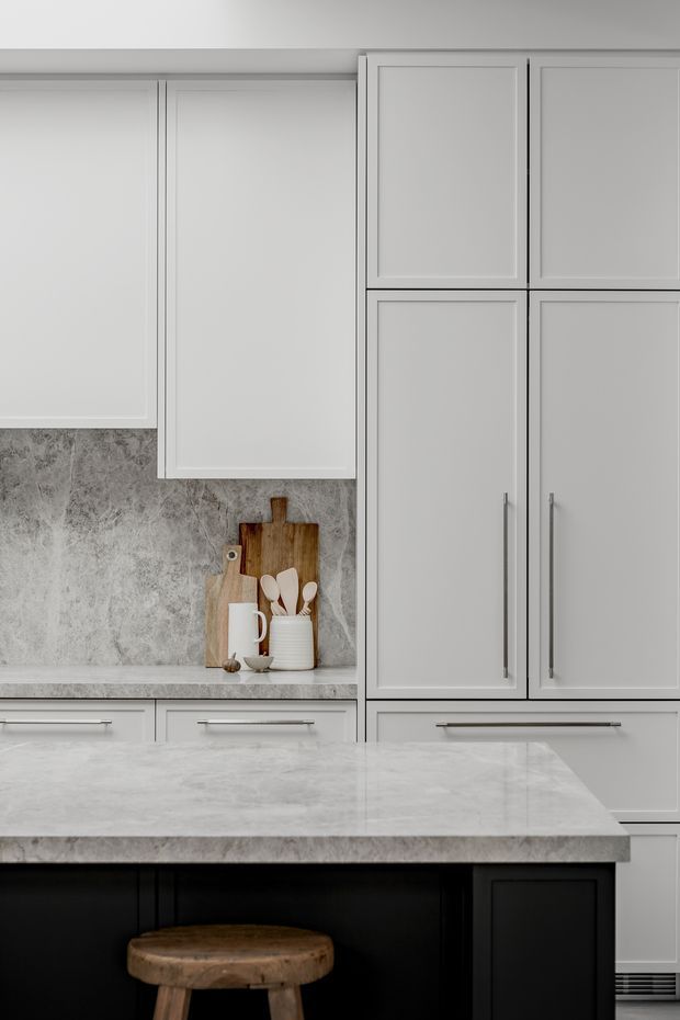 Close-up view of a refined kitchen featuring light grey shaker-style cabinetry, a mottled grey stone-look splashback and benchtop, brushed metal handles, and a dark island base with a wooden stool. A small styled niche contains cutting boards, crockery, and utensils.