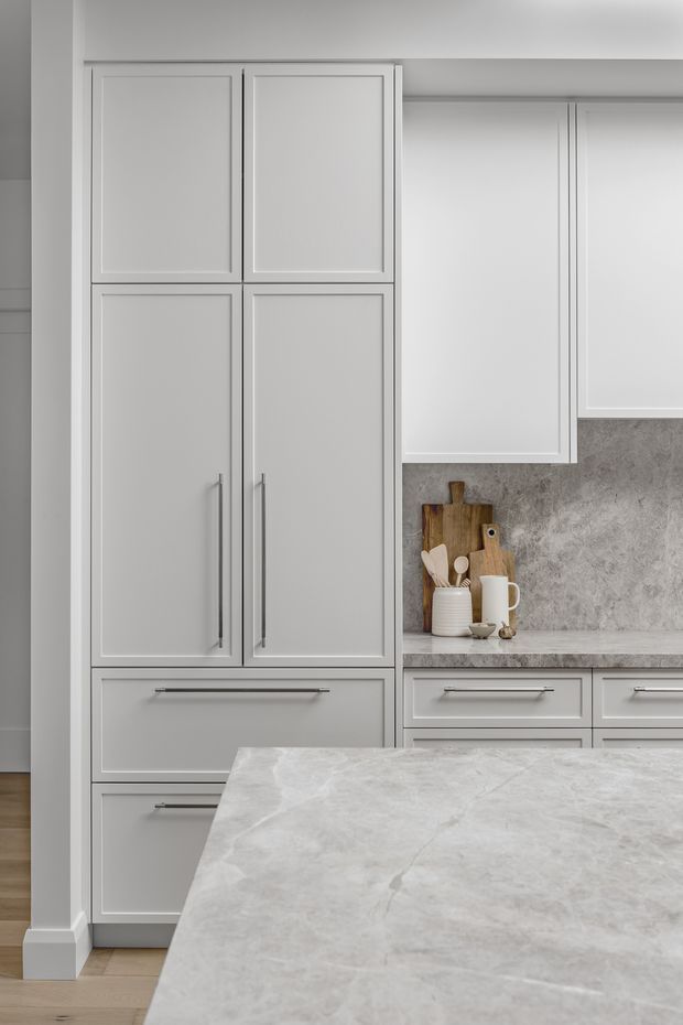 A close-up view of a refined, light-toned kitchen featuring tall Shaker-style cabinetry, lower drawers, a pale grey stone island benchtop in the foreground, matching stone benchtops, and a textured grey stone splashback. Minimal countertop styling includes wooden boards, utensils, and a ceramic mug.