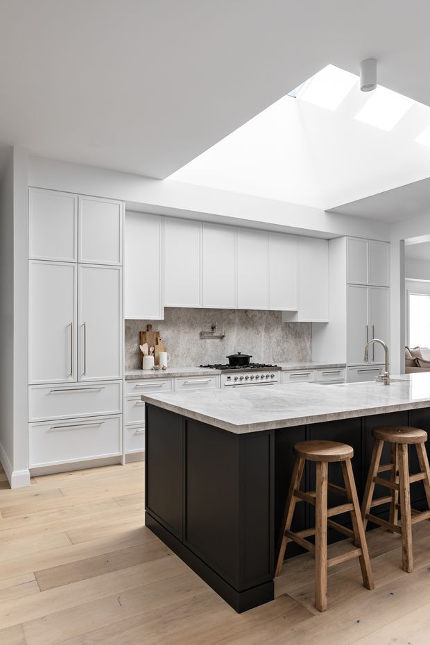Bright, refined kitchen with extensive white Shaker-style cabinetry, a dark island base, pale stone benchtops, a textured stone splashback, timber flooring, and seating for two at the island. A large ceiling skylight floods the room with natural light, while a living area is partially visible through the opening at right.