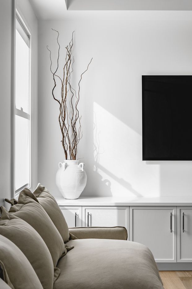 Bright, refined living room corner with a large upholstered sectional sofa in the foreground, white built-in cabinetry beneath a wall-mounted television, and a white decorative vase filled with tall branches. Strong daylight enters through the tall side window, creating crisp shadows on the white wall. Light timber flooring and a restrained neutral palette give the space a clean, contemporary feel.