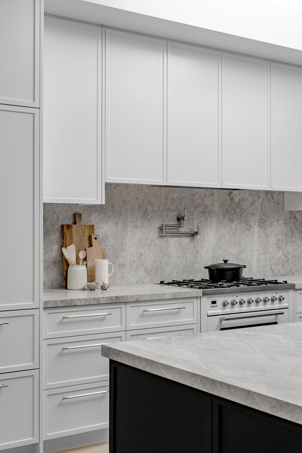 A refined, monochromatic kitchen with tall white Shaker-style cabinetry, extensive grey veined stone-look splashback and benchtops, a stainless steel range cooker, and a dark island with a matching pale countertop. Wooden cutting boards and neutral ceramic utensils add subtle warmth.