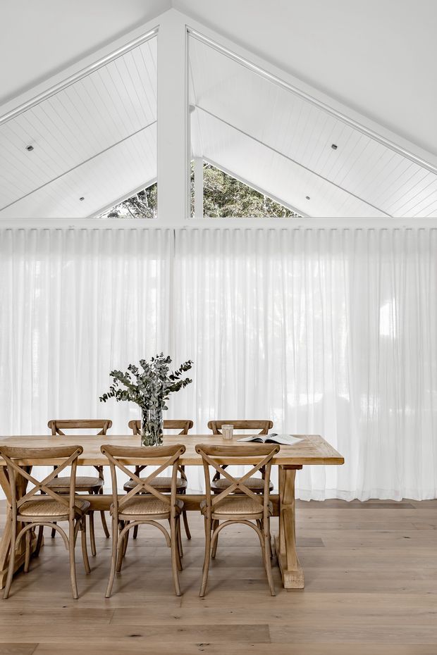 Bright dining room with a long rustic timber dining table and matching cross-back chairs, set against sheer white curtains. The room features pale timber flooring and a dramatic white pitched, panelled ceiling with triangular clerestory-style glazing and recessed downlights, creating an airy, light-filled interior.