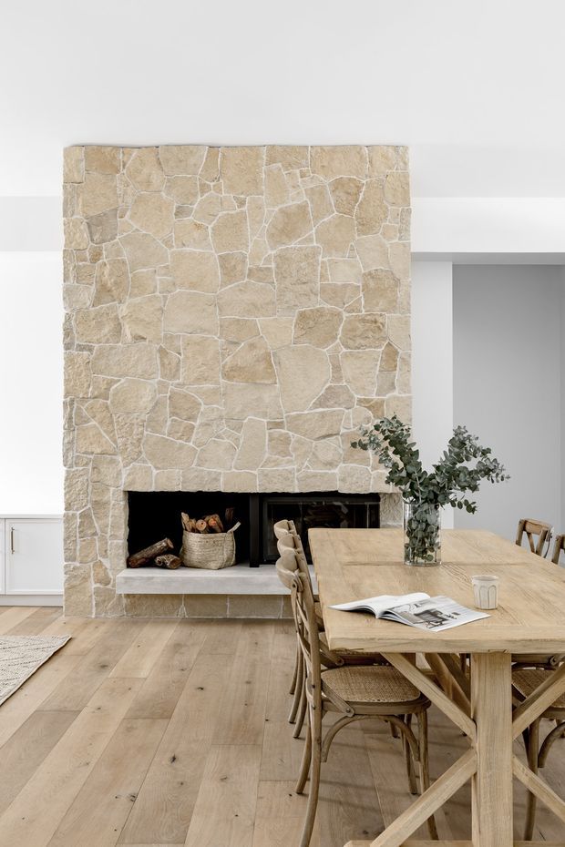 Bright open-plan dining and living area featuring a large rustic irregular-stone fireplace surround with a dark firebox, pale wide-plank timber flooring, white walls and cabinetry, and a substantial light timber dining table with woven wooden chairs. The styling is warm, natural, and understated.