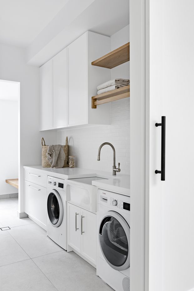 Bright, compact contemporary laundry room with a white built-in cabinetry run, white front-loading washer and dryer, pale grey benchtop, white subway-tile splashback, and open timber shelving holding folded towels. A deep brushed-metal gooseneck tap sits over a utility sink. The space has large light-grey floor tiles and a black vertical door pull, creating a clean, highly functional fit-out.