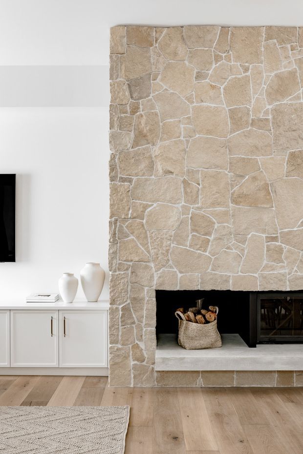 A bright contemporary living room centred on a large floor-to-ceiling irregular stone fireplace with a wide concrete hearth and black firebox. White built-in cabinetry with simple dark pulls sits to the left beneath a wall-mounted television, while pale timber flooring, neutral ceramics, and a textured light rug create a calm, warm interior.