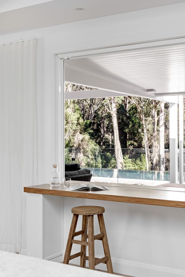 Bright white open-plan interior with a timber breakfast bar and rustic timber stool in the foreground. The space opens through large glazing to a covered outdoor area, swimming pool and wooded landscape. White curtains, wall panelling, a slatted ceiling and clean-lined detailing create an airy, refined coastal feel.