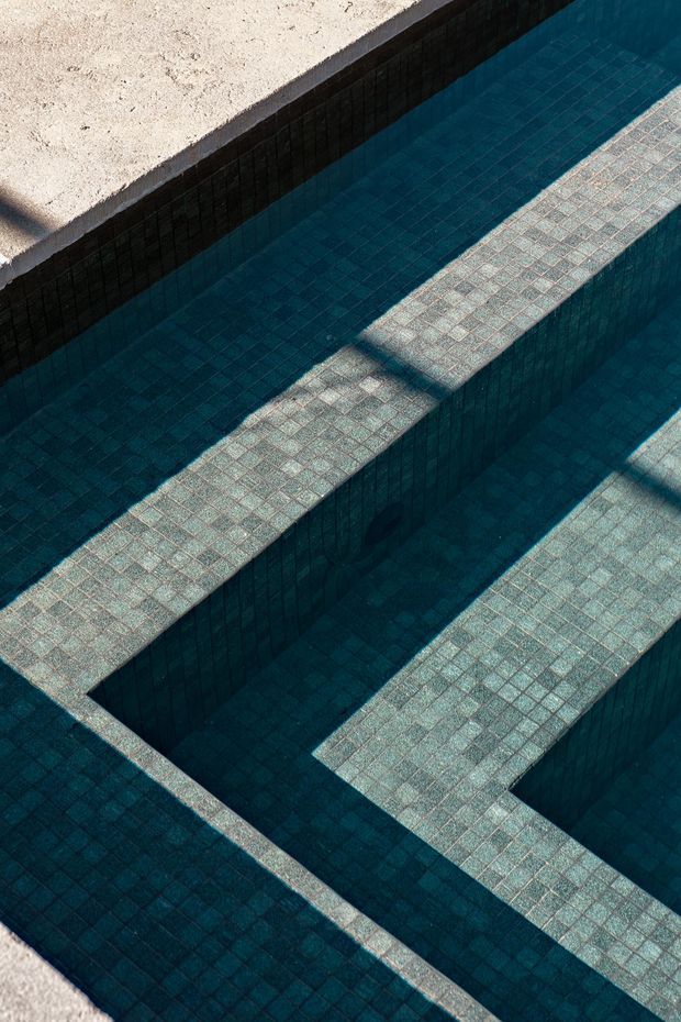 Abstract close-up architectural photograph of a tiled swimming pool, showing deep blue-green mosaic tiles, stepped or partitioned submerged platforms, and strong geometric shadows. A pale textured pool surround is visible along the upper and lower edges.
