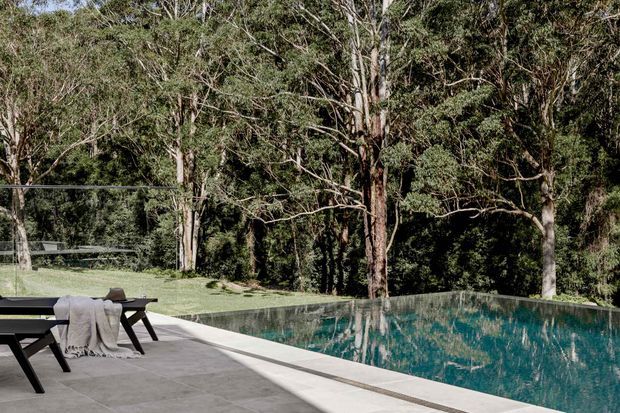 A tranquil outdoor pool area set within a wooded landscape. A rectangular swimming pool with a pale paved surround occupies the foreground, while dark outdoor dining furniture and a draped throw sit at the poolside. Mature trees and dense vegetation form a private natural backdrop.