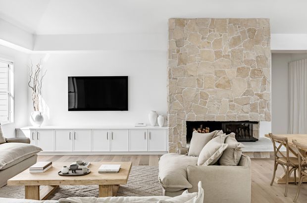 Bright, refined open-plan living room with a large pale natural-stone fireplace surround, white built-in media cabinetry, wall-mounted television, neutral upholstered seating, timber coffee table, and an adjacent dining area. The space has abundant natural light, light timber flooring, and a calm neutral palette.