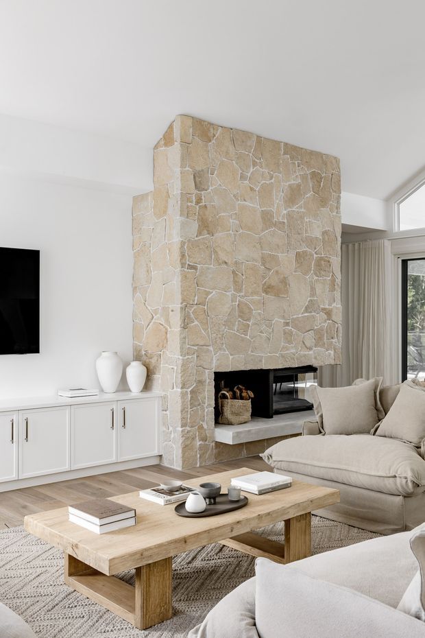 Bright, refined living room with a large irregular light-stone fireplace surround as the focal point. The room includes a white built-in media cabinet, wall-mounted television, neutral upholstered sofas, a substantial natural timber coffee table, pale patterned rug, and floor-to-ceiling windows with sheer curtains.