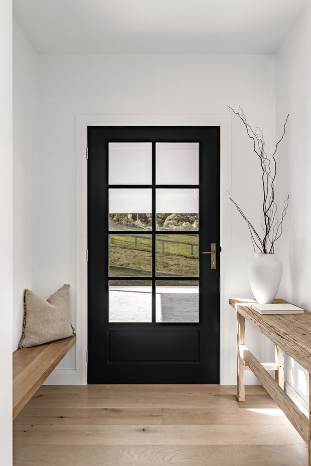 A bright, minimalist entryway with white walls, pale timber flooring, a black glazed panel door, and simple rustic timber furnishings. A built-in bench with a neutral cushion sits to the left, while a rough timber console table with a white ceramic vase and branches is positioned to the right. The door looks onto a grassy outdoor area and paved terrace.