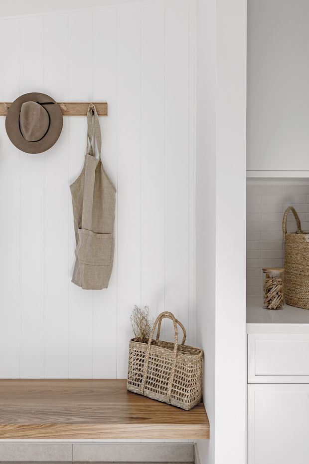A bright, softly styled entryway or mudroom nook with white vertical wall panelling, a warm timber bench, and a simple timber wall-mounted hook rail. A neutral linen apron and woven accessories add texture, while white built-in cabinetry and open shelving with baskets are visible to the right. The overall look is calm, natural, and minimalist with coastal influence.