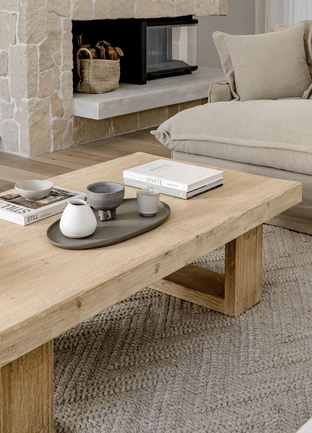 A warm, neutral living room centred on a substantial light timber coffee table with chunky legs and a lower stretcher. A textured grey-beige rug, upholstered linen-toned armchair and sofa, ceramic tableware, and stacked books create a relaxed, refined setting. A stone-clad fireplace with a black firebox and concrete hearth forms the architectural backdrop.