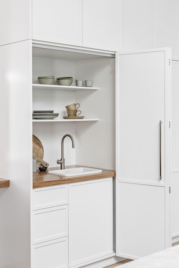 A bright, compact built-in butler's pantry or kitchenette with white shaker-style cabinetry, open display shelving, a timber benchtop, undermount sink, and brushed metal gooseneck tap. The cabinetry includes a tall concealed storage cupboard with a long vertical handle, while neutral ceramic tableware and timber boards sit on the shelves. The space has a clean, refined, warm-neutral aesthetic.