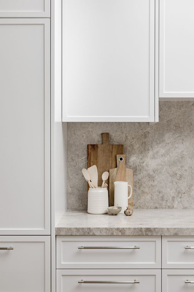 Close-up view of a refined kitchen run featuring pale grey-white shaker-style cabinetry with recessed panel doors and slim metallic bar pulls. A light stone-look benchtop and matching textured stone splashback frame a small styling arrangement of timber cutting boards, a ceramic utensil crock, a white jug, and a small bowl. The composition is bright, calm, and softly neutral with traditional detailing.