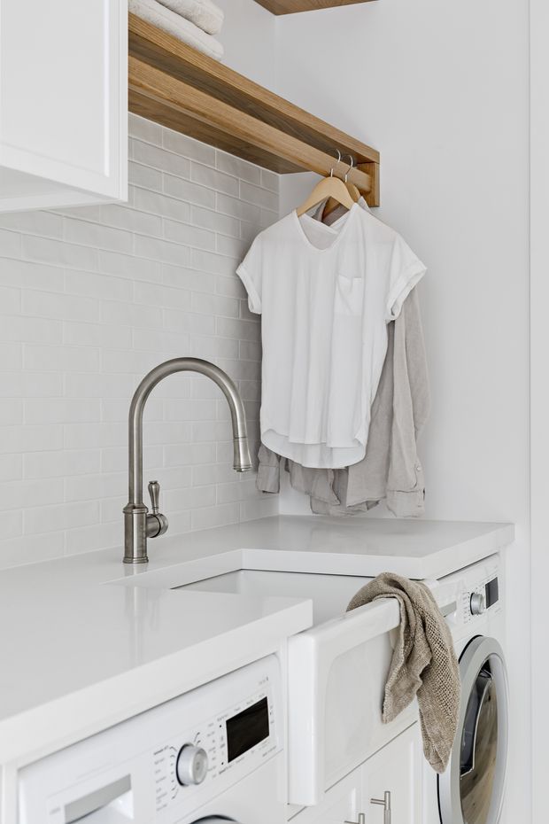 Bright, compact laundry room with white cabinetry and appliances, a white tiled subway backsplash, pale engineered stone benchtops, and warm timber open shelving with hanging rails. A stainless steel gooseneck tap sits above the laundry sink, with front-loading washing appliances below and folded towels and clothing adding context.