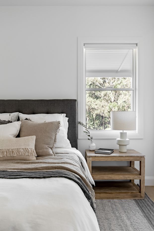 A calm, softly lit bedroom with a partially visible upholstered bed, layered white and beige bedding, fringed throws, and neutral cushions. A rustic timber bedside table with open shelves holds a ceramic lamp and small decor. White walls, a white-framed window, and a grey woven rug create a restrained neutral scheme, with leafy trees visible outside.