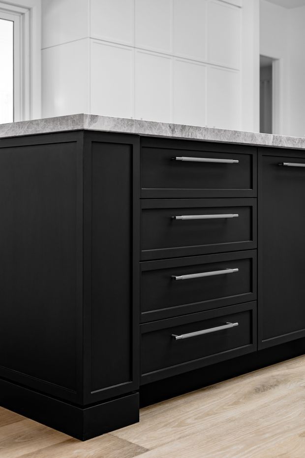 Close-up interior view of a contemporary kitchen island or base cabinetry, featuring matte black shaker-style doors and drawers with long silver bar handles, a pale grey veined stone benchtop, white wall panelling, and light timber flooring.