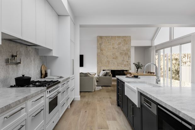 A bright open-plan kitchen opening into a living area. The kitchen features white shaker-style cabinetry, a large pale stone island with a farmhouse sink, dark island base cabinetry, a freestanding range, and a textured stone backsplash. Light timber flooring continues into the living room, which has a substantial natural-stone fireplace surround. Floor-to-ceiling glazing brings in abundant daylight.