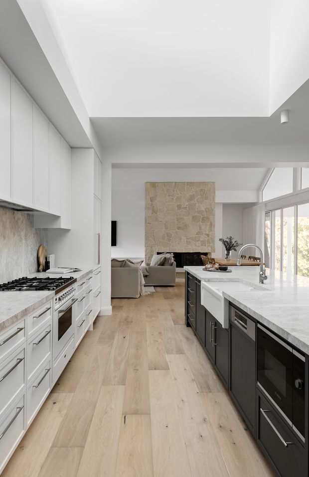 A bright, spacious contemporary kitchen viewed along a central passage toward an adjoining living area. The kitchen features white shaker-style cabinetry, a large pale stone island with an integrated farmhouse sink, dark lower island cabinetry, stainless-steel appliances, and wide light timber flooring. A stone fireplace feature wall and extensive glazing are visible in the living area beyond.