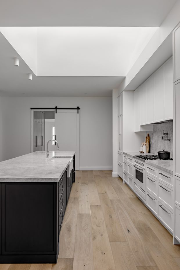 A bright, refined kitchen with a large dark island, pale stone benchtop, white Shaker-style cabinetry, integrated range and cooktop, grey stone splashback, and light timber flooring. A large skylight or clerestory opening brings in abundant natural light, while a black-framed sliding door is visible at the rear.