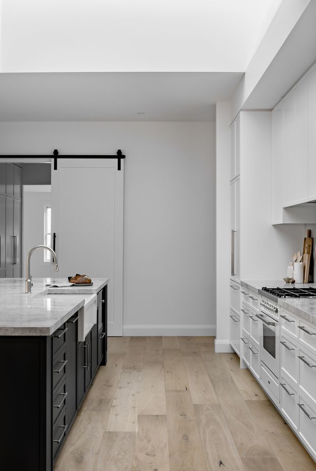 A bright, refined galley-style kitchen with a large central island. The room features white shaker-style cabinetry, dark island cabinetry, pale stone countertops, integrated appliances, a gas cooktop, stainless hardware, and wide-plank light timber flooring. A black-framed barn-style sliding door is visible at the rear, with predominantly white walls and cabinetry creating a clean, spacious appearance.
