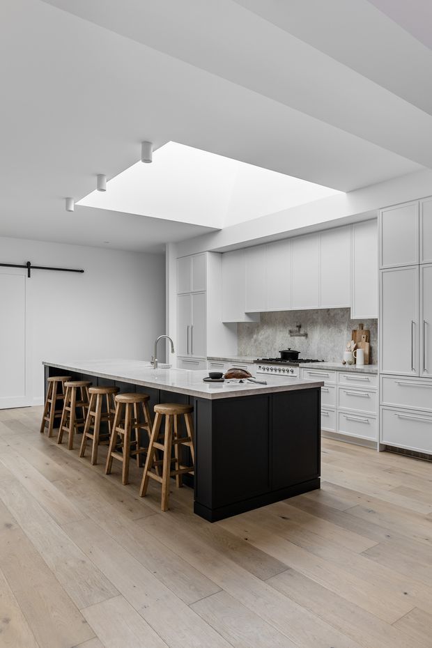 A bright, refined kitchen with a large white stone island and black cabinetry base, seating for multiple people on wooden stools, extensive white shaker-style cabinetry, a pale stone backsplash, integrated appliances, and light timber flooring. A large skylight or ceiling opening provides abundant natural light, while a barn-style sliding door is visible at left.