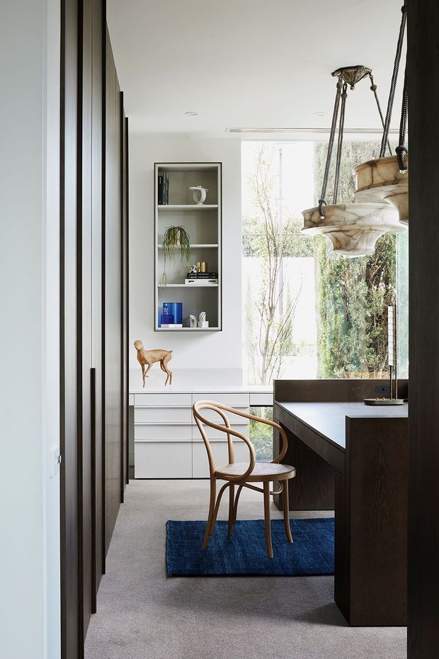 A refined home office viewed through a doorway, featuring a dark timber built-in desk and cabinetry, a light upholstered chair with curved timber arms, grey carpet, and a blue area rug. White walls contain a recessed display shelf with books and objects, while large windows provide abundant natural light and views of greenery. Sculptural suspended pendant lights hang above the workspace.
