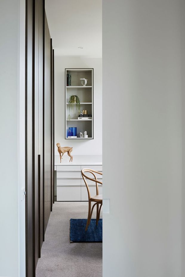 A narrow view through a partially open doorway into a refined, minimalist interior. The room features a white built-in credenza with multiple drawers, a recessed open display shelf, pale carpet, and a light timber dining chair partly visible over a small deep-blue rug. Dark timber-framed doors or wall panelling line the left side, while a plain light wall frames the right side. Decorative objects and a plant occupy the shelving, but the image is primarily about the passage and glimpse into the furnished room.