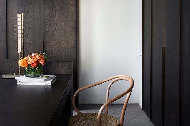 A refined, dark-toned interior corner featuring a black built-in console or desk, full-height dark timber wall panelling and cabinetry, a white wall, grey carpet, and a sculptural woven-seat timber chair. A vase of orange flowers and stacked books sit on the console, adding a warm accent to the otherwise moody, minimalist composition.