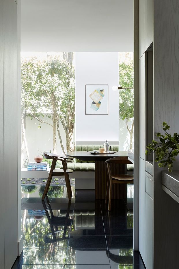 A bright, refined dining nook viewed through a doorway or narrow passage. The space features a round dark dining table with sculptural wooden chairs, a built-in upholstered banquette in muted sage green, large floor-to-ceiling glazing opening to leafy planting, and a framed abstract artwork. Glossy black tiled flooring strongly reflects the furniture and greenery. Pale vertical wall panelling and dark built-in joinery frame the view, with a slim wall-mounted light beside the artwork.