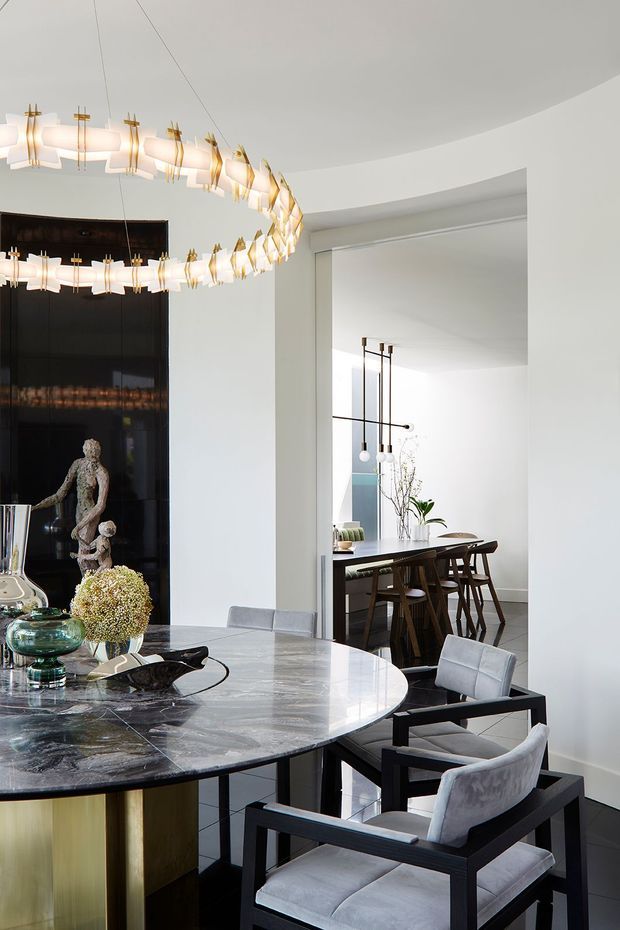 Elegant contemporary dining room with a large round dark marble dining table featuring a metallic gold base, upholstered black-framed armchairs, and a sculptural ring pendant light with white illuminated elements and gold accents. White walls and a curved ceiling create a refined setting, while a black feature wall with artwork and an open view toward a secondary dining or living area add depth.