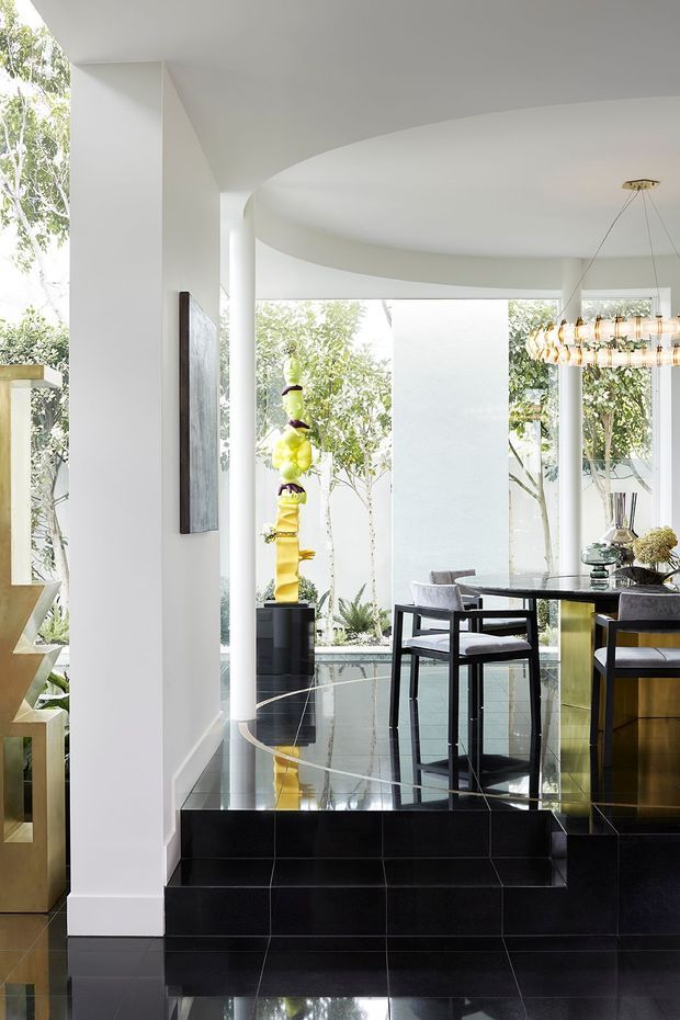A bright, high-end open-plan interior with a dining area elevated above a black tiled floor. The space features a dramatic curved white ceiling, extensive floor-to-ceiling glazing, black reflective flooring and steps, sculptural artwork, a glass dining table with dark-framed chairs, a statement pendant light, and dark built-in cabinetry with a warm metallic-gold finish. Lush greenery is visible outside through the glazing.