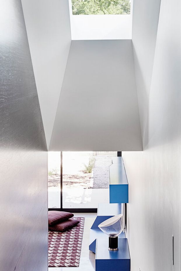 A narrow, bright modern interior viewed down a stairway or passage, with crisp white walls and a sculptural angular ceiling void. At the lower level, floor-to-ceiling glazing opens toward an outdoor area, while blue built-in cabinetry, a patterned rug, cushions, and a small decorative table arrangement add colour and contrast.