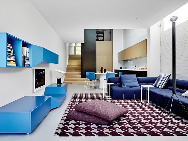 Bright contemporary open-plan living room with a bold blue and white palette. The space features built-in blue shelving and low media cabinetry, a deep blue sectional sofa, white tubular side tables and chairs, a patterned burgundy-and-white area rug with floor cushions, and a compact kitchen area beyond. A timber staircase rises at the rear, while a black vertical fireplace or heater and white brick-textured wall add contrast.