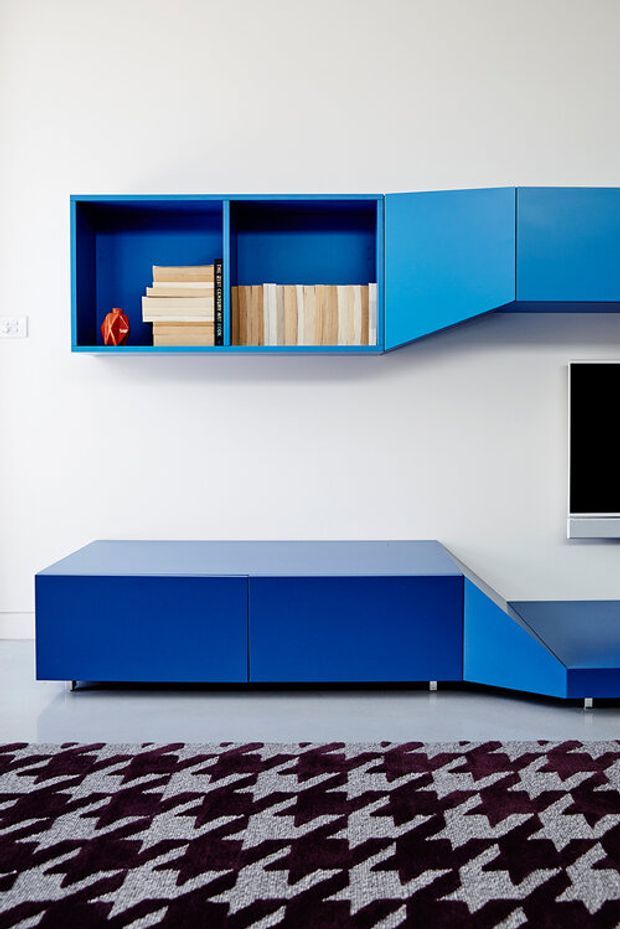 A highly stylised contemporary living room media wall featuring vivid blue built-in cabinetry. Open wall shelves display books and timber samples, while a low blue media console connects to an angled side unit beneath a partially visible television. White walls and a patterned burgundy, black and light-grey rug provide contrast.