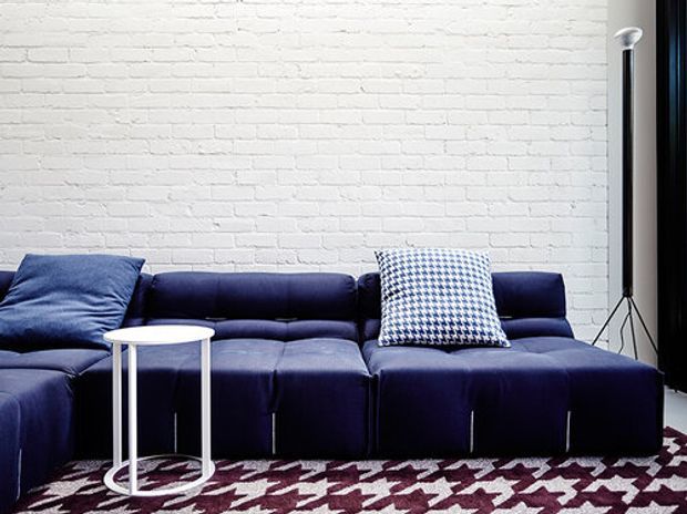 A contemporary living room featuring a large deep navy modular sectional sofa, a small white round side table, a patterned navy-and-white cushion, and a bold burgundy and white patterned rug. The room has a white painted exposed-brick wall, a black floor lamp, and dark window or curtain elements.