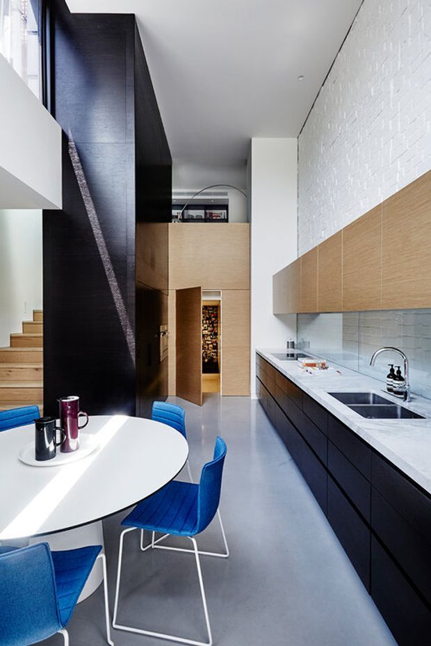 A tall, narrow contemporary kitchen and dining area with a dramatic double-height volume. The space features dark timber cabinetry and a long white benchtop, pale timber wall cabinetry, white-painted brick walls, polished grey flooring, a sculptural dark stair enclosure, and bright blue dining chairs around a white oval table. Strong natural light enters from the left, creating a crisp, high-contrast interior.