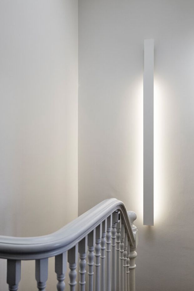 A minimalist interior stairway with a curved, traditional white-painted balustrade and turned spindles. Smooth light-grey walls surround a tall, narrow vertical wall light that casts a soft glow, creating a refined and quiet atmosphere.