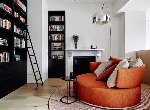 A refined living room with a large curved burnt-orange sectional sofa, pale textured cushions, a round rug and slim black side table. Black built-in floor-to-ceiling bookshelves with a rolling ladder line the left wall, while a traditional white mantel fireplace and black firebox anchor the rear wall. A metallic pendant light hangs above, and light timber flooring adds warmth to the predominantly white and black interior.