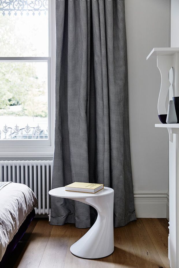 A refined bedroom corner featuring a tall window with a decorative upper grille, full-length black-and-white patterned curtains, a white radiator, timber flooring, and a sculptural white side table beside the edge of a bed. White architectural trim and shelving contribute to a classic, monochromatic interior.