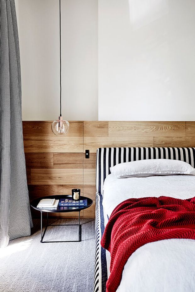 A bright, contemporary bedroom featuring a bed with a bold black-and-white striped upholstered headboard, white bedding, and a red textured throw. The room has a warm timber-panelled feature wall, pale carpet, a small black side table, a clear globe pendant light, and full-height patterned curtains.