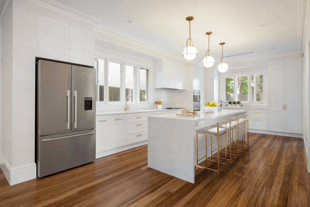 Bright, spacious white kitchen with a large central island, integrated white cabinetry, stainless-steel French-door refrigerator, timber flooring, and three globe pendant lights. Tall windows and glazed doors provide abundant natural light, while brass-toned bar stools and hardware add subtle warmth.