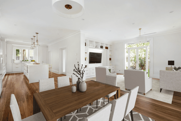 Bright open-plan interior combining a white kitchen, dining area, and living room. The space features warm timber flooring, a dark timber dining table with upholstered white chairs, white shaker-style cabinetry and built-in media joinery, large windows and glazed doors, recessed lighting, and restrained contemporary furnishings.